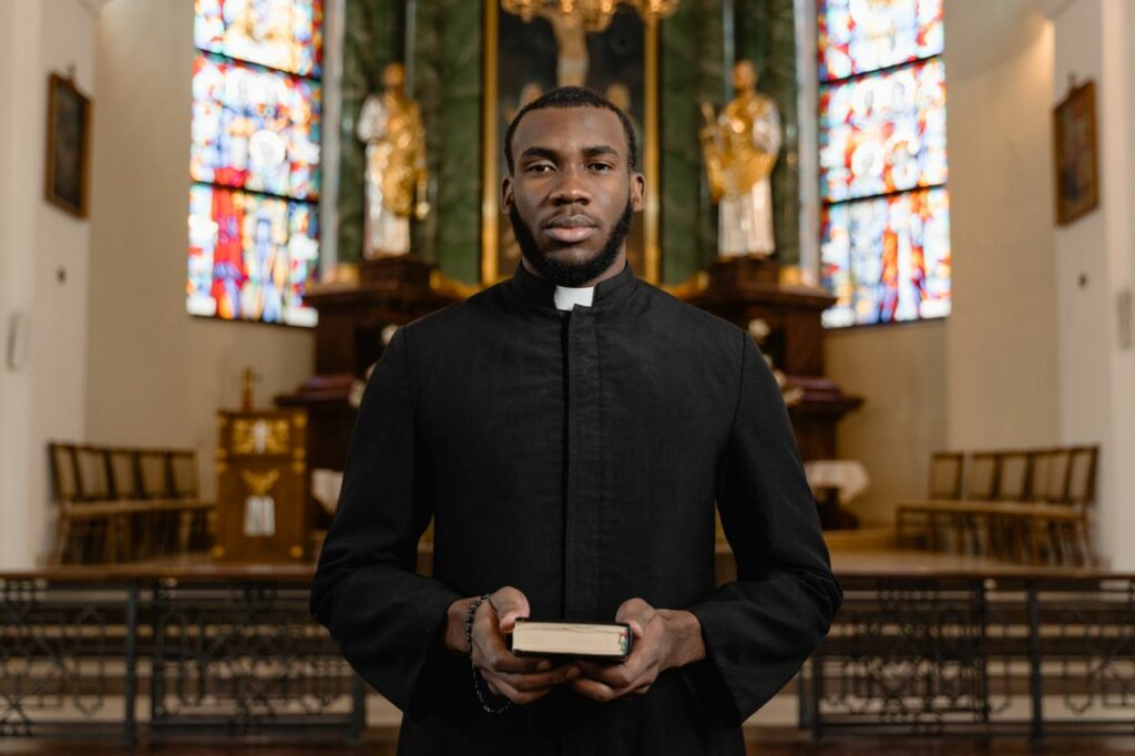 Portrait of a priest in a cassock holding a Bible in an ornate church interior.