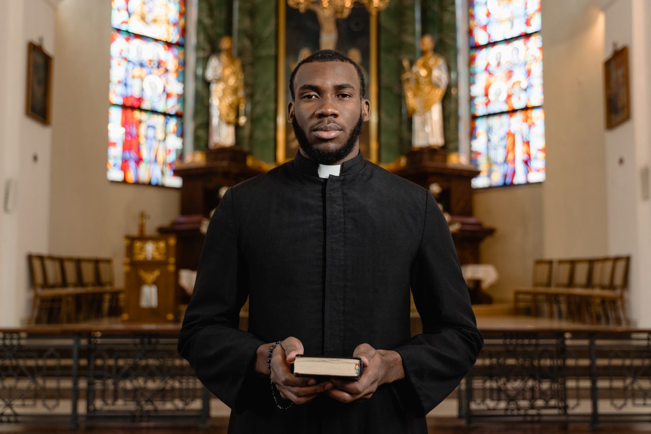 Portrait of a priest in a cassock holding a Bible in an ornate church interior.