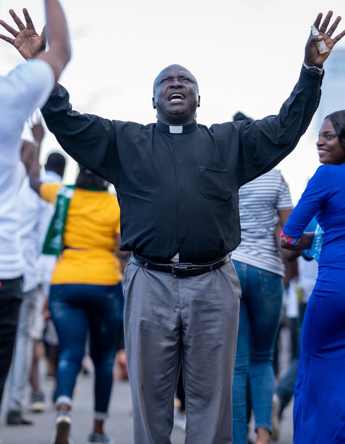 African priest with arms raised singing joyfully in a public gathering outdoors.
