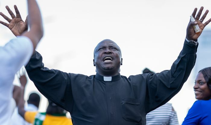 African priest with arms raised singing joyfully in a public gathering outdoors.